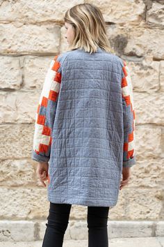 a woman standing in front of a stone wall wearing an orange and blue jacket with stripes on the sleeves