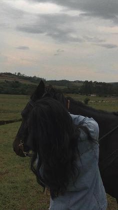 a woman standing next to a black horse on top of a lush green field under a cloudy sky