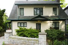 a white house with green trim and black shutters on the front door is next to a brick fence