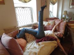 a woman laying on top of a couch covered in pillows next to a table and window