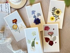 four cards with pressed flowers on them are laying next to an open book and hat