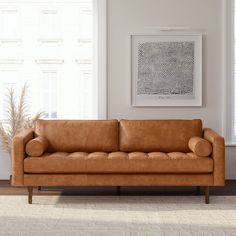 a tan leather couch sitting in a living room next to a white rug and two windows