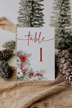 a table number card with pine cones and evergreens on the side, sitting next to each other