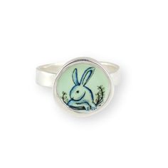 This adorable Rabbit ring has a vintage storybook quality.  Available in whole sizes 6 through 9 I carved the sterling silver setting from a block of wax, then cast that wax pendant into metal and molded it so I can make multiples. Both sides of the setting then get a layer of vitreous enamel (fine glass) and fired at 1400 degrees in a kiln, four separate times, to build strength and color saturation . Finally I take an illustration from my sketchbook, turn it into an enamel screen print, and fire that on the top for a durable, waterproof and vibrant finish. So many steps. I ship this in a gift box using First Class Mail with tracking. All jewelry and photographs copyright of Mark Poulin Whimsical Sterling Silver Rings For Gifts, Silver Whimsical Rings For Gifts, Whimsical Silver Rings For Gifts, Engraved Enamel Ring As A Gift, Whimsical Sterling Silver Ring As Gift, Whimsical Silver Rings For Gift, Hand Painted Silver Jewelry For Anniversary, Whimsical Sterling Silver Ring Gift, Whimsical Round Sterling Silver Jewelry