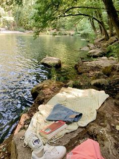 an open book and some clothes are on the edge of a river with trees in the background