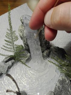 a hand is holding a piece of glass with water coming out of it and some plants in the background