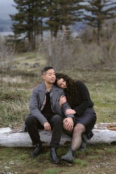 a man and woman sitting on a log in the woods, one is hugging the other