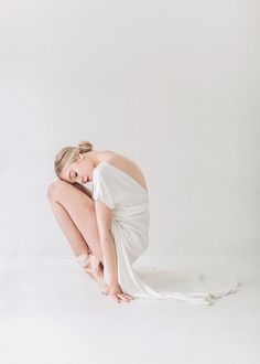 a woman kneeling on the ground with her head down and hands behind her back, wearing a white dress