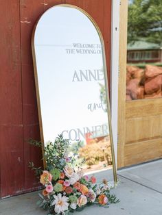 a large mirror sitting on the side of a building next to flowers and greenery