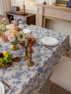 a blue and white table cloth with flowers, candles, plates and fruit on it