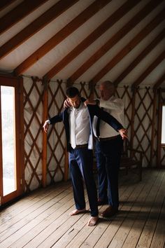 two men standing in a room with wooden floors and walls, one wearing a white shirt and the other a blue suit