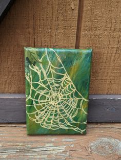 a green and yellow painting on wood with spider web in the center, hanging on a wooden wall