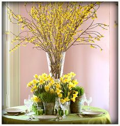 yellow flowers in vases on a table with plates and wine glasses, near a pink wall