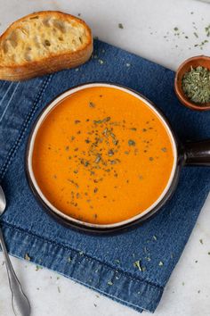 a bowl of carrot soup with bread and herbs on the side next to spoons