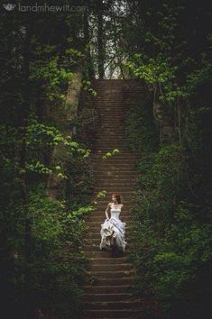 a woman in white dress sitting on steps surrounded by trees and greenery, with her back turned to the camera