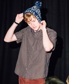 a young man wearing a blue hat while standing in front of a black curtain with his hands on his head