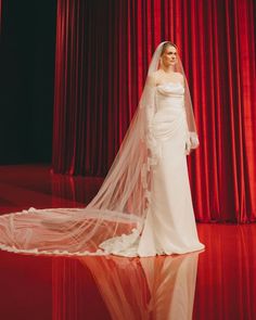 a woman in a white wedding dress and veil standing on a red stage with curtains behind her