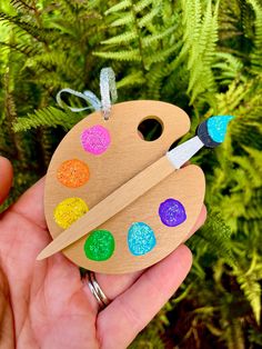 a hand holding a wooden ornament with paintbrushes and dots on it