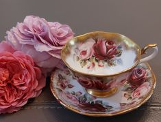 two pink flowers sitting next to a tea cup and saucer on top of a table