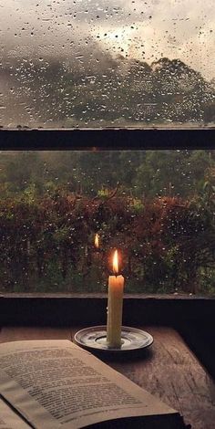 a lit candle on a plate next to an open book and window with raindrops