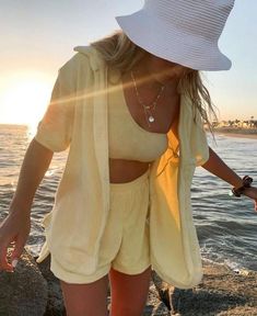 a woman in yellow shorts and a white hat is walking along the beach at sunset