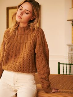 a woman sitting on top of a wooden table wearing a brown sweater and white pants