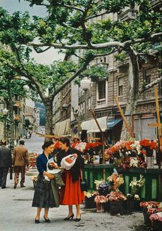 two women standing in the middle of an open air market with flowers and fruit on display