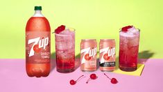 three bottles and two glasses filled with soda on a pink counter top next to cherries