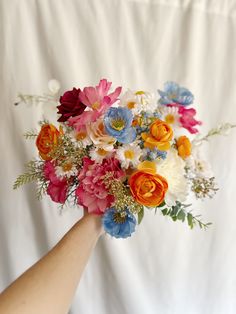 a person holding a bouquet of flowers in front of a white background with an orange, pink and blue flower arrangement