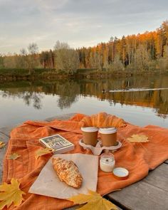 two cups of coffee sit on an orange blanket next to a lake with autumn leaves