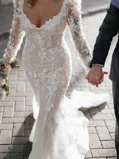 a bride and groom holding hands walking down the street in their wedding gowns with long sleeves