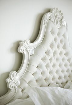 a white bed with an ornate headboard and pillow on it's side, in front of a white wall