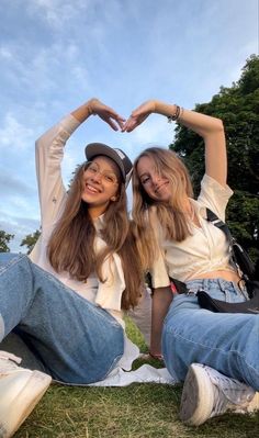 two young women sitting on the ground making a heart shape with their hands while wearing jeans and baseball caps