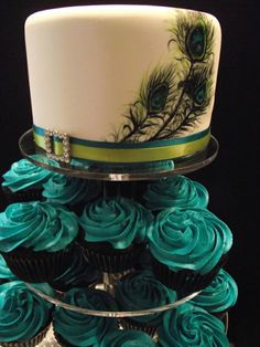 two tiered cake with blue frosting and peacock feathers on the top, surrounded by cupcakes