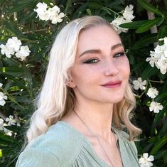 a woman with blonde hair standing in front of white flowers and greenery, looking at the camera