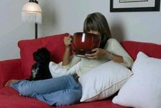 a woman sitting on a red couch holding a coffee cup and a black poodle