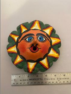 an orange and green sun face with blue eyes next to a measuring ruler on a white surface