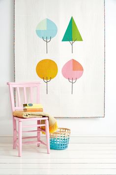 a chair next to a wall hanging with colorful trees on it and a basket in the foreground