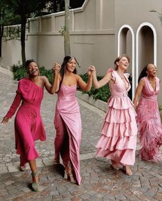 four women in pink dresses are dancing on a cobblestone sidewalk with their arms around each other