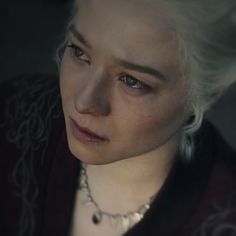 a close up of a person with white hair wearing a black jacket and pearls necklace