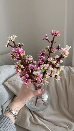 a person holding a glass vase with flowers in it on a couch next to a pillow