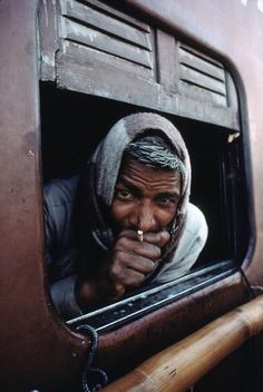 a man looking out the window of a train with his hand on his nose and wearing a head scarf