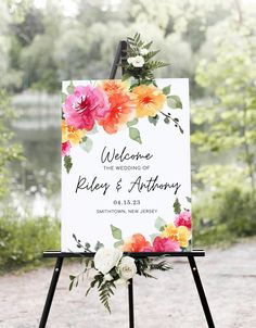 a welcome sign with flowers on it in front of some trees and water at an outdoor wedding