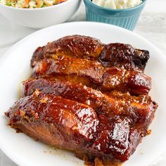 ribs covered in bbq sauce on a white plate next to bowls of coleslaw