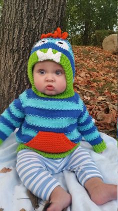 a baby wearing a knitted frog outfit sitting in front of a tree