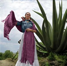 a woman is standing in front of a large cactus and wearing a purple dress with her arms outstretched