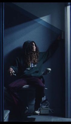 a man with long hair sitting on a chair in a dark room holding his arms out