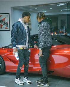 two young men standing next to a red sports car in a showroom, talking