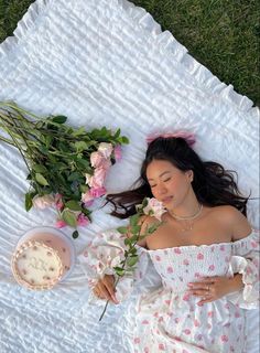 a woman laying on top of a white blanket next to flowers and a bowl of cake