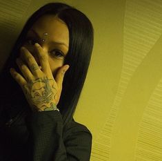 a woman with tattoos covering her face and hand over her mouth, standing against a yellow wall